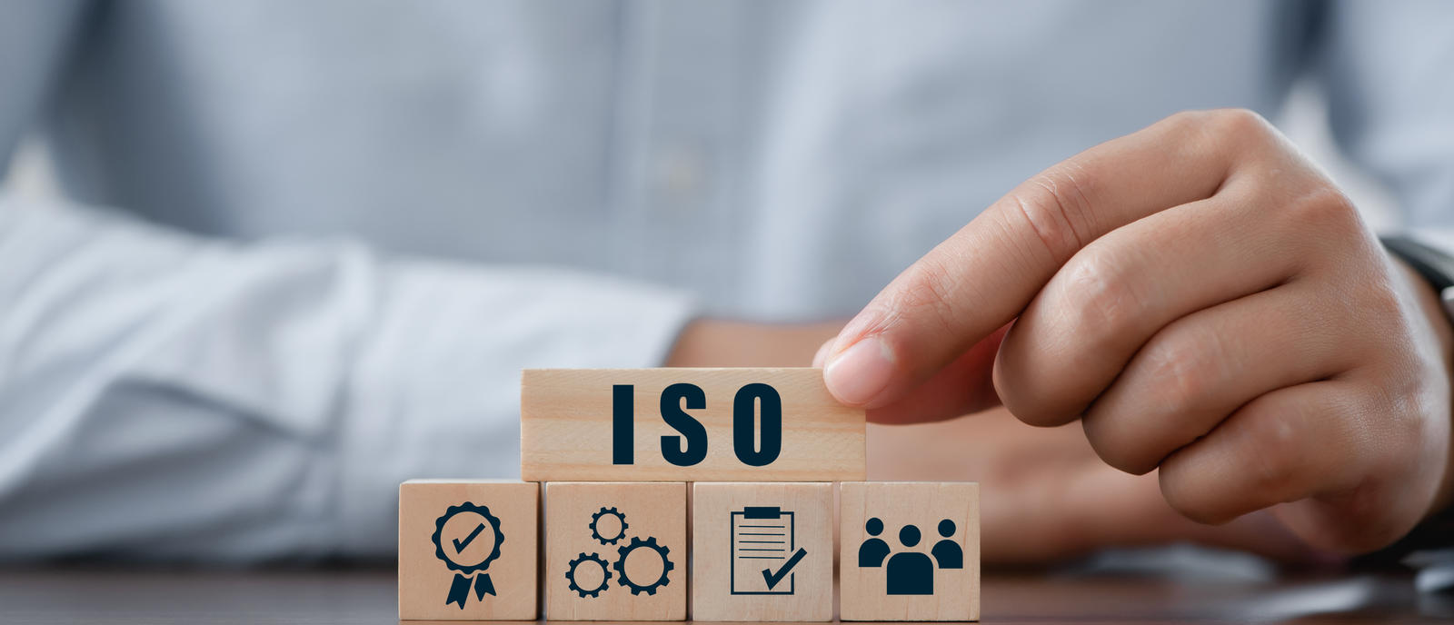 Eine Person platziert einen Holzblock mit der Aufschrift ‚ISO‘ auf weitere Blöcke, die verschiedene Aspekte eines Managementsystems symbolisieren