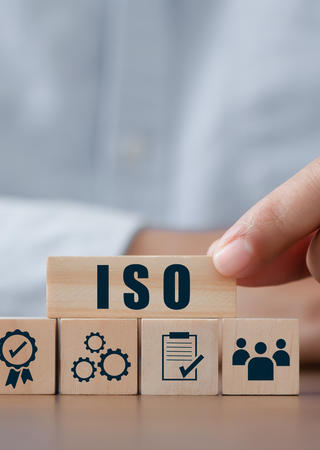 Eine Person platziert einen Holzblock mit der Aufschrift ‚ISO‘ auf weitere Blöcke, die verschiedene Aspekte eines Managementsystems symbolisieren